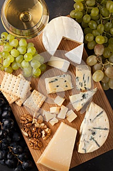 Assorted cheeses with white grapes, walnuts, crackers and white wine on a wooden Board. Food for a romantic date on a dark stone b