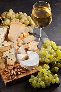 Assorted cheeses with white grapes, walnuts, crackers and white wine on a wooden Board. Food for a romantic date on a dark stone b