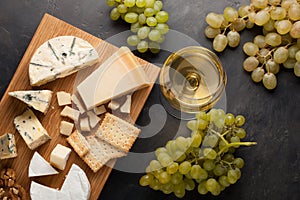 Assorted cheeses with white grapes, walnuts, crackers and white wine on a wooden Board. Food for a romantic date on a dark stone b
