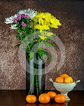 Assorted Chrysanthemum Flowers in a Glass Vase with Chalice Bowl and Satsuma Oranges on a Wooden Table Top
