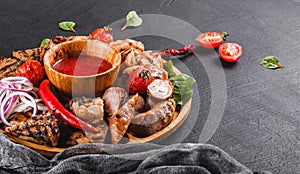 Assorted delicious grilled meat and sausages with tomatoes and bbq sauce on cutting board over black stone background. Hot Meat