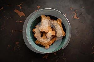 Assorted dog biscuits in a teal bowl on a dark textured surface, top view, suitable for pet food advertising, dog