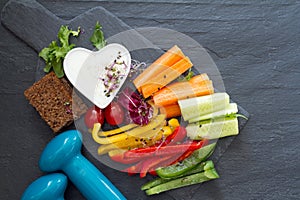 Assorted fresh chopped vegetables with dip and dumbbells on dark background, sport and healthy food concept