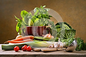 Assorted fresh vegetables with chopping board, ladle and old crock pot in rustic setting on a wooden table.