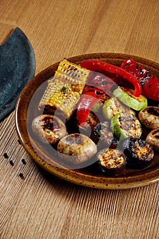 Assorted grilled vegetables - mushrooms, zucchini, peppers, corn in a ceramic plate on a wooden background. Vegetarian warm salad