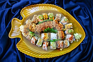 Assorted sushi rolls arranged in a yellow fish platter with blue scrunched fabric background