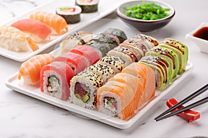 Assorted sushi rolls on a white square dish, seaweed salad and chopsticks on a white stone