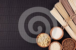 Assortment of bundles raw noodles with ingredient in wooden bowls on black striped mat background with copy space, top view.