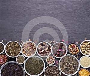 Assortment of dried tea leaves, fruit and herbs in bowls on dark, stone background