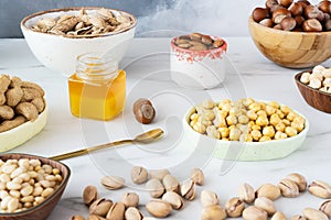 An assortment of nuts hazelnuts, almonds, cashews, pine nuts and chickpeas in different bowls are on a white table.