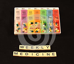 An assortment of pills in a colorful weekly pill box above weekly medicine spelled in tiles on a black background