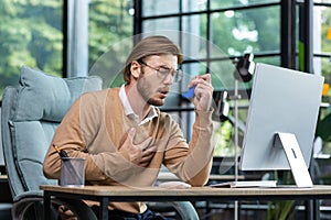 Asthma attack, panic attack. A young man sits in the office at the table, holds his chest, blows an inhaler in his mouth