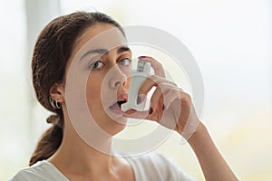Asthma. Portrait of a young woman holding an inhaler to her mouth and looking at the camera. Blurry background. Copy