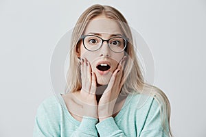 Astonished blonde female model with spectacles keeps mouth widely opened, looks amazed, keeps hands on cheek, remembers