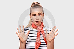 Astonished blonde woman gestures stop sign with both hands, screaming loudly, wearing striped t-shirt and red scarf, posing