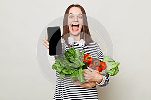 Astonished excited brown haired young woman embraces bouquet of fresh vegetables wearing striped casual shirt isolated over gray
