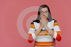 Astonished lady with dark hair covers mouth with both hands, looks in disbelief at camera, wears spectacles and casual jumper,