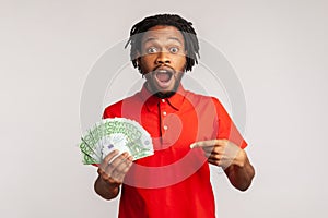 Astonished man with open mouth wearing red casual style T-shirt, hold lot of money, pointing at euro