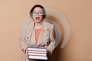 Astonished woman school teacher holds heavy volumes of hardcover books, expresses stupefaction at camera, cream backdrop