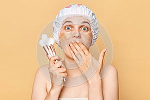 Astonished surprised woman doing makeup holding powder brushes posing with patches under eyes isolated over beige background