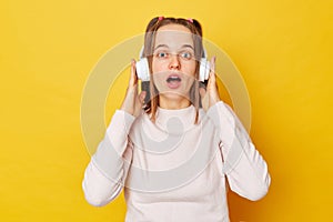 Astonished teen girl wearing jumper and headphones posing isolated over yellow background staring bugged eyes having startled