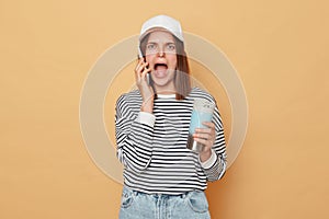 Astonished woman wearing baseball cap and striped shirt holding thermos and using phone isolated over beige background hearing bad