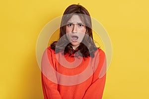 Astonished woman with widely opened mouth posing  over yellow background  wearing orange jumper  sees something shocking