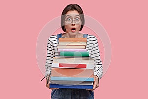 Astounded young European woman with dark hair, dressed in striped clothes, carries many books, being stupefied to learn