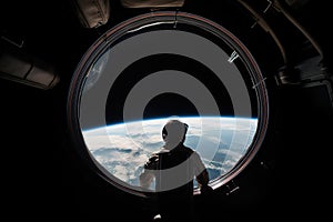 Astronaut stand near round window with view on earth in space station in space