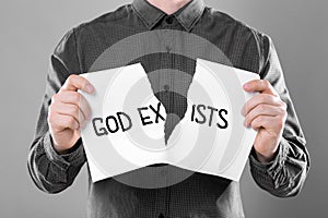 Atheism as religious position. Man ripping sheet of paper with text God Exists on grey background, closeup