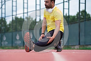 Athlete in activewear and headphones stretches leg, on running track , warming up before training
