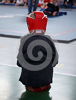 A young athlete boy in boxing gloves and a protective headgear looks at the tatami with the fighters.