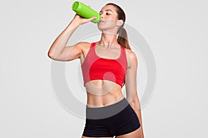 Athlete female drinks water, being thirsty after jogging during sunny day, has slim body, dressed in casual top and shorts, poses