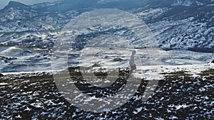 An athlete jogs along a snow-covered trail at the top of a hill against the backdrop of a mountain range. Aerial view