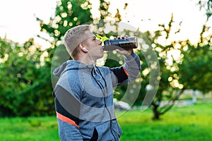 Athlete man is drinking water protein. A bodybuilder listens to music while training. In the summer in the park in the