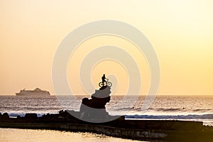 Athlete with mountain bike over a big cliff for success concept with ocean and cruise boat with tourists in background . sunset