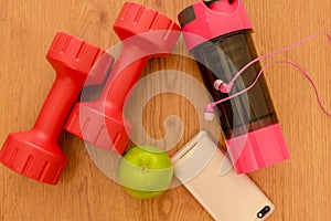 Athlete`s set with two pink dumbbells, smarphone with pink headphones, green appple and bottle of water on wooden