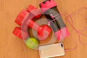 Athlete`s set with two pink dumbbells, smarphone with pink headphones, green appple and bottle of water on wooden