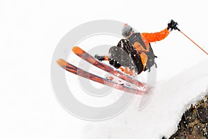 The athlete skier on a light background with a jump moves off the roof of a snow-covered hut with flying flakes of snow