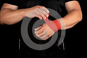 athlete stand in black clothes and wrap his hands in red textile elastic bandage