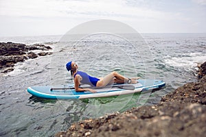 athlete woman in a blue swimsuit and bandana on lies paddle board on a quiet blue ocean. Sup surfing in water