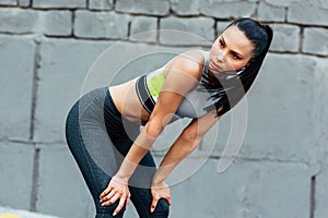 Athlete young woman resting next to the gray concrete wall after workout outdoor and looking to the camera. Fitness girl resting