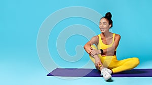 athletic african american woman sitting on yoga mat, blue background