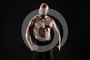 Athletic bald, bearded, tattooed man in black shorts is posing against a black background. Close-up portrait.