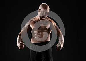 Athletic bald, bearded, tattooed man in black shorts is posing against a black background. Close-up portrait.