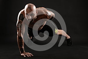 Athletic bald, tattooed man in black shorts and sneakers is posing against a black background. Close-up portrait.