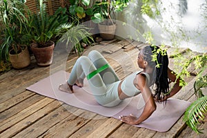 Athletic black woman do exercises with fitness elastic band sitting on sported mat in home greenery