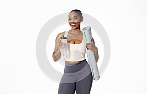 Athletic black woman holds water bottle and fitness mat, studio