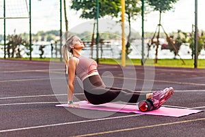 Athletic blond sporty woman training on mat outdoor summer day, massaging and stretching hamstring and ankle leg muscles