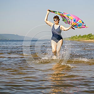 Athletic elderly woman runs along the waves on a summer sunny day against the background of mountains